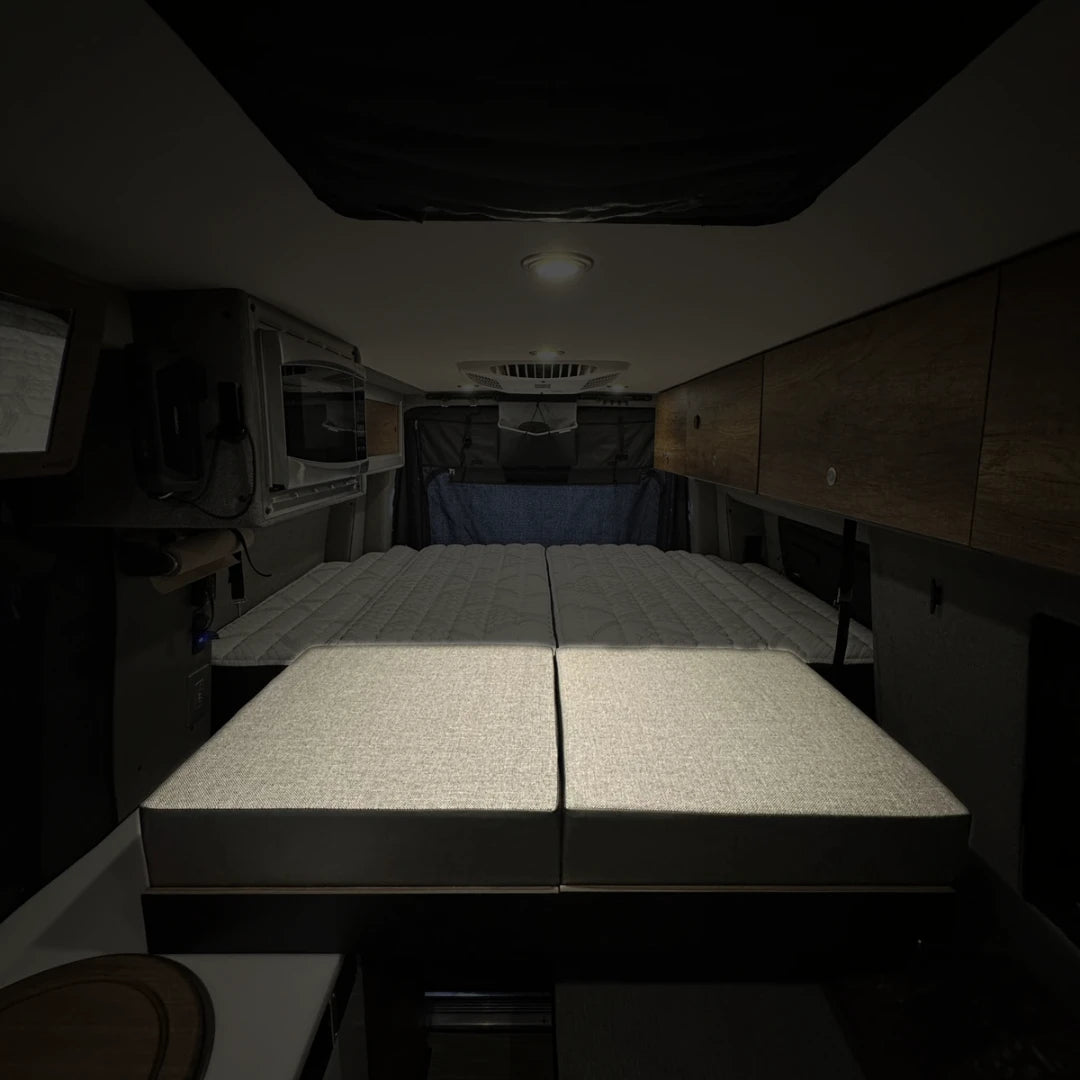 Storyteller Overland Mode van bed system with extended cushions and overhead cabinets in dim lighting