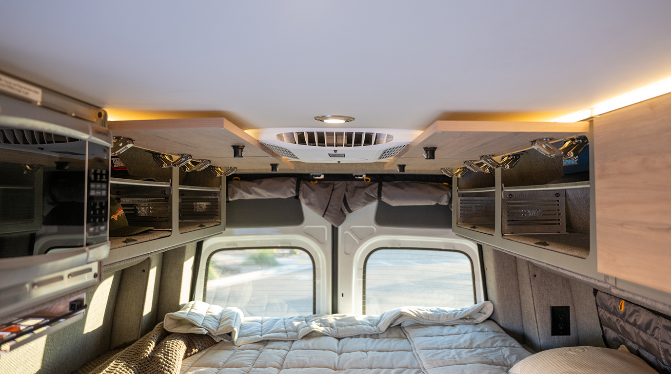 Interior van view showing overhead Storyteller cabinet shelves above a bed with windows in the rear doors.