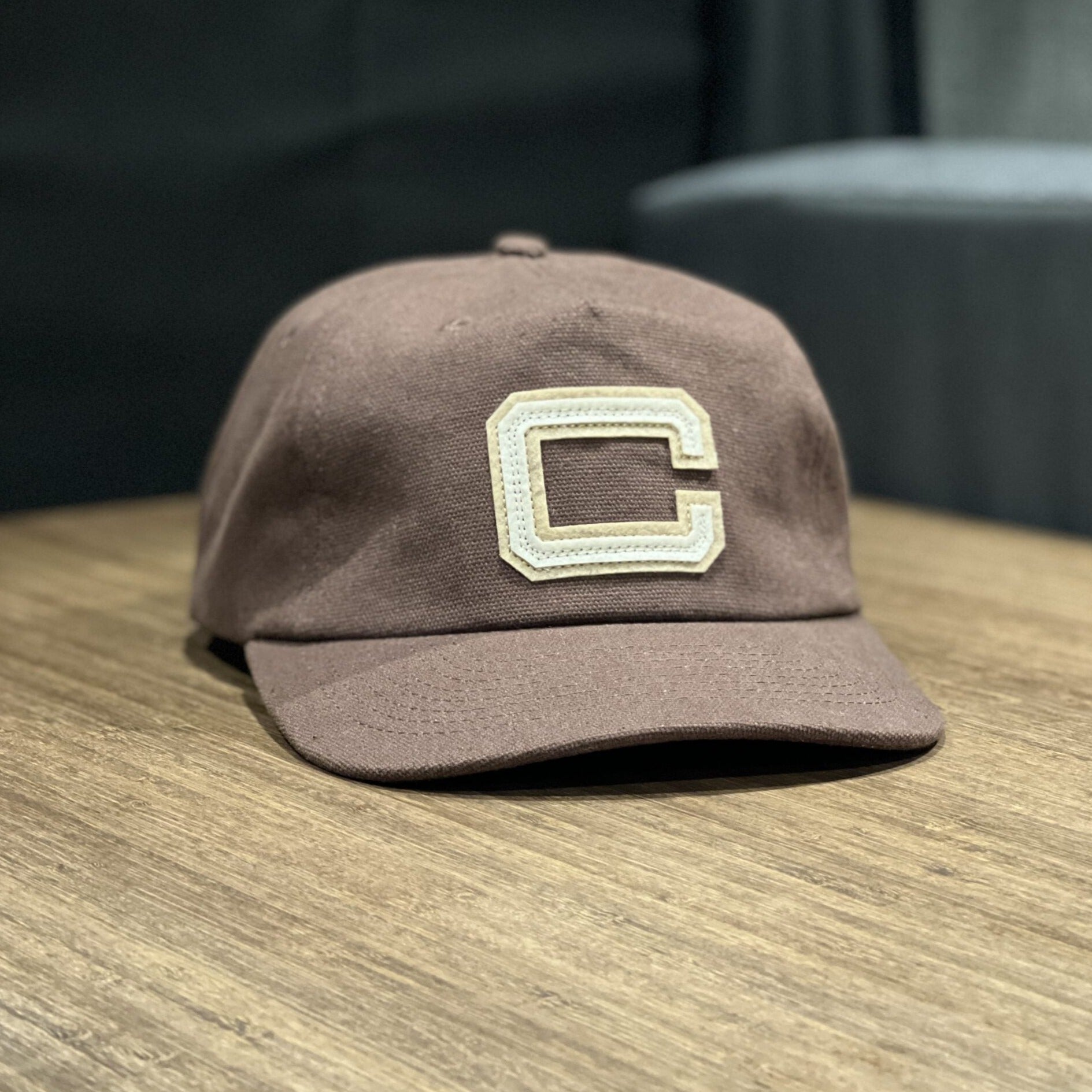 Brown Revel Letterman Hat with embroidered cream C on front, displayed on wooden table