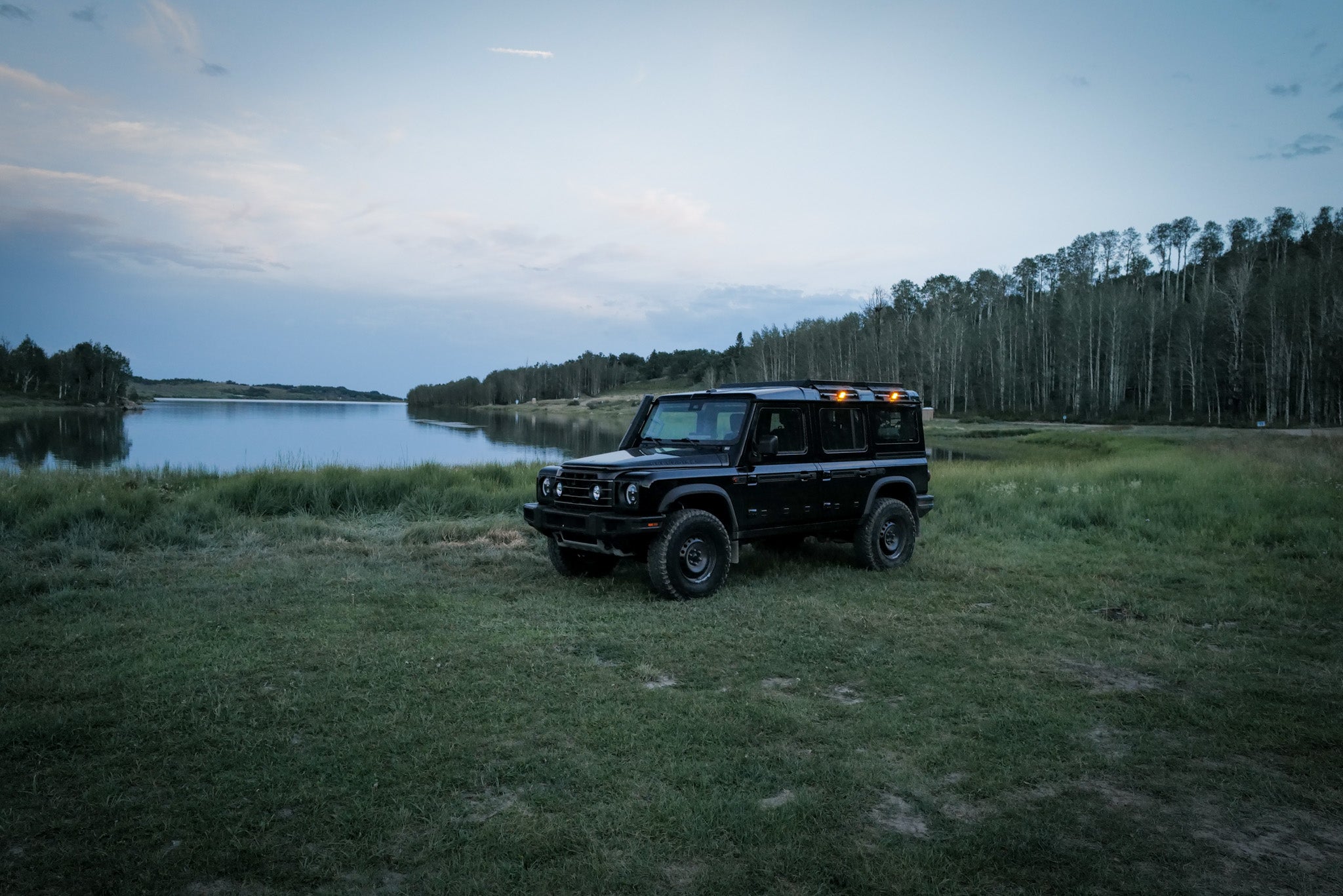 A black off-road INEOS Grenadier parked on a grassy field next to a calm lake, with a line of trees in the background under a twilight sky.