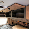 Open Storyteller overhead cabinet shelf in van interior with wood finish and black compartments.