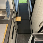 Folded black floor mattress laid out in center aisle of van interior with kitchenette and seating area.